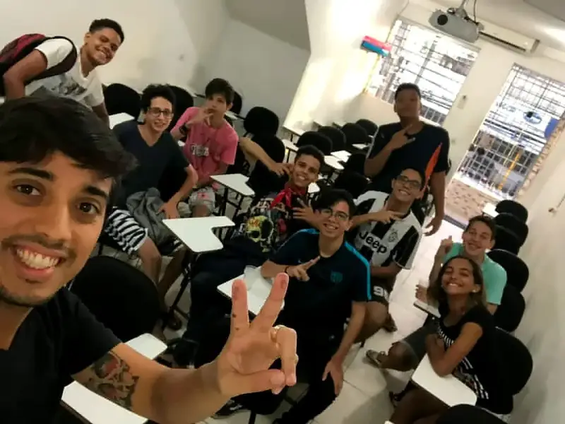 Selfie de um aluno fazendo sinal de paz, com a turma sorrindo nas carteiras